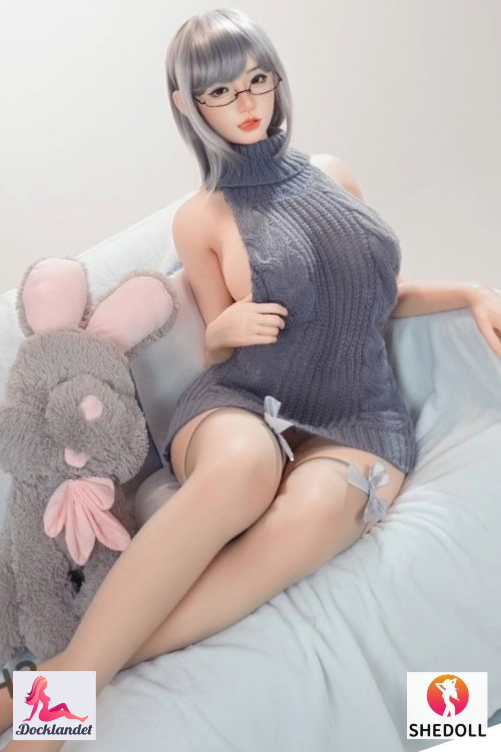 Shedoll sexdocka med kort grå frisyr och glasögon i grå stickad klänning sittandes i soffa bredvid kanin-gosedjur – realistisk silikon-docka från Docklandet
